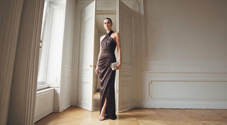 Elegant woman in a dark satin evening gown stands in a bright, classic room with tall windows, paneled walls, and herringbone floors.
