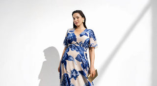 Woman in a blue and white floral dress holding a gold clutch against a plain white background with shadow lines.