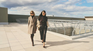 Two stylish women walk across a sunlit rooftop terrace, one in a beige coat and sunglasses, the other in black, with modern railings and a cloudy sky.