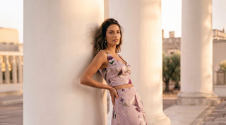 A woman in a floral purple outfit leans against a white column in a sunlit, classical setting. Her expression is calm and confident.