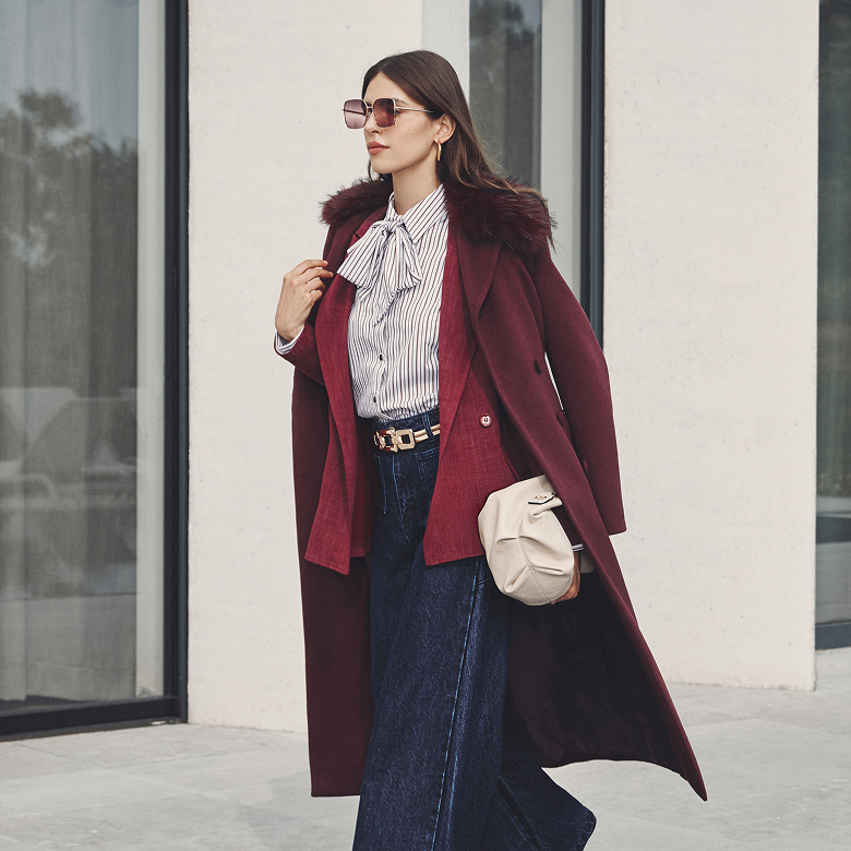 Donna in un cappotto bordeaux e pantaloni larghi, in piedi con un'espressione serena