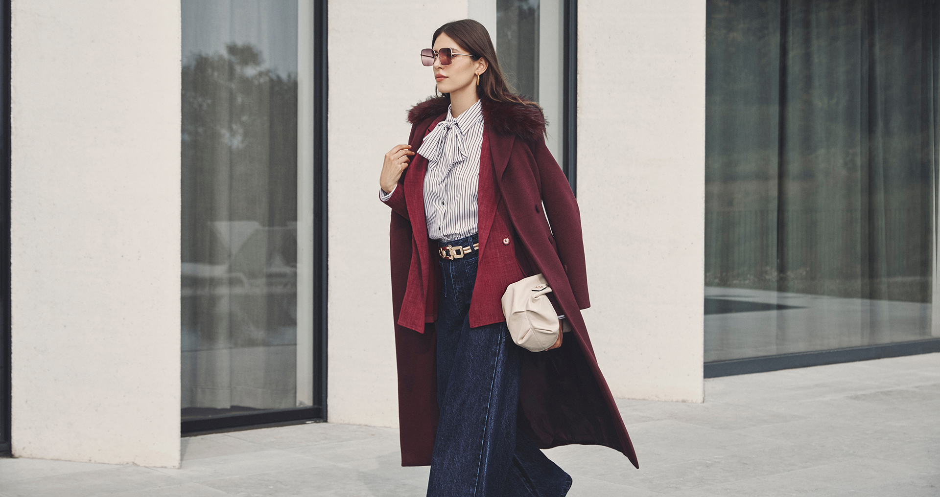 Donna in un cappotto bordeaux e pantaloni larghi, in piedi con un'espressione serena