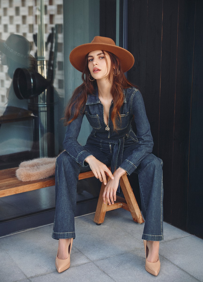Donna con cappello e tuta di jeans seduta su una panchina in un ambiente all'aperto