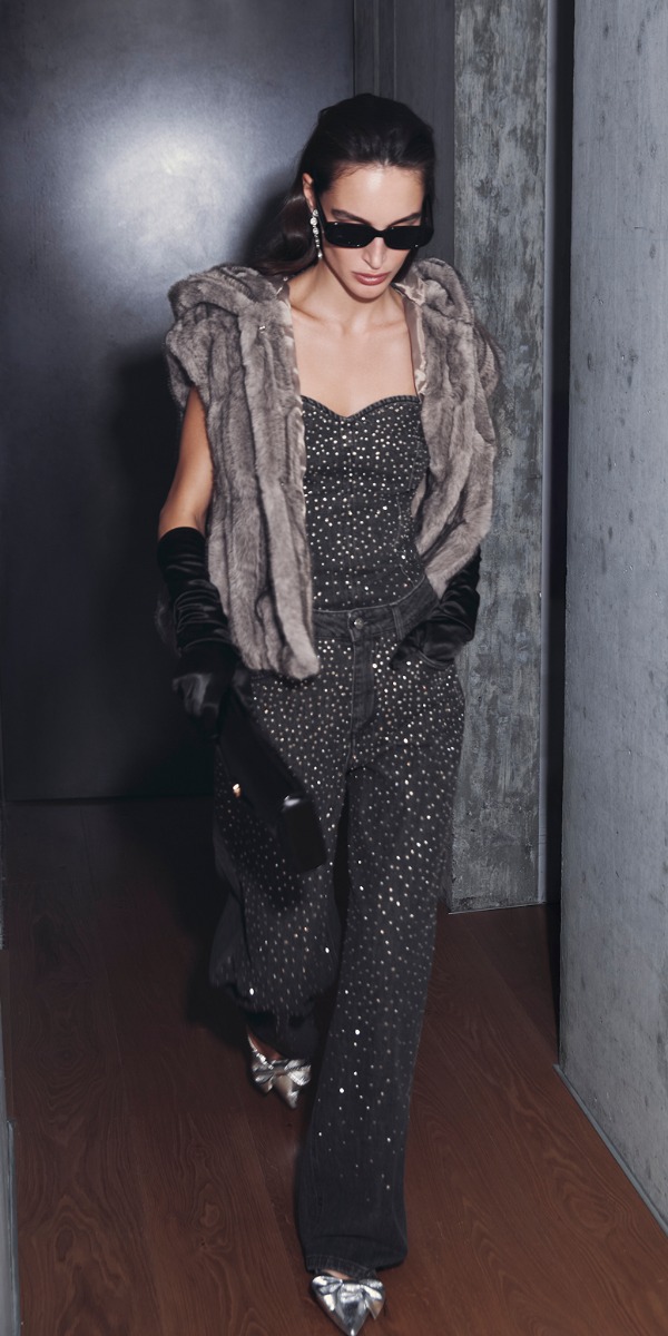 Donna in tuta nera con gilet di pelliccia, posa elegante in un ambiente urbano