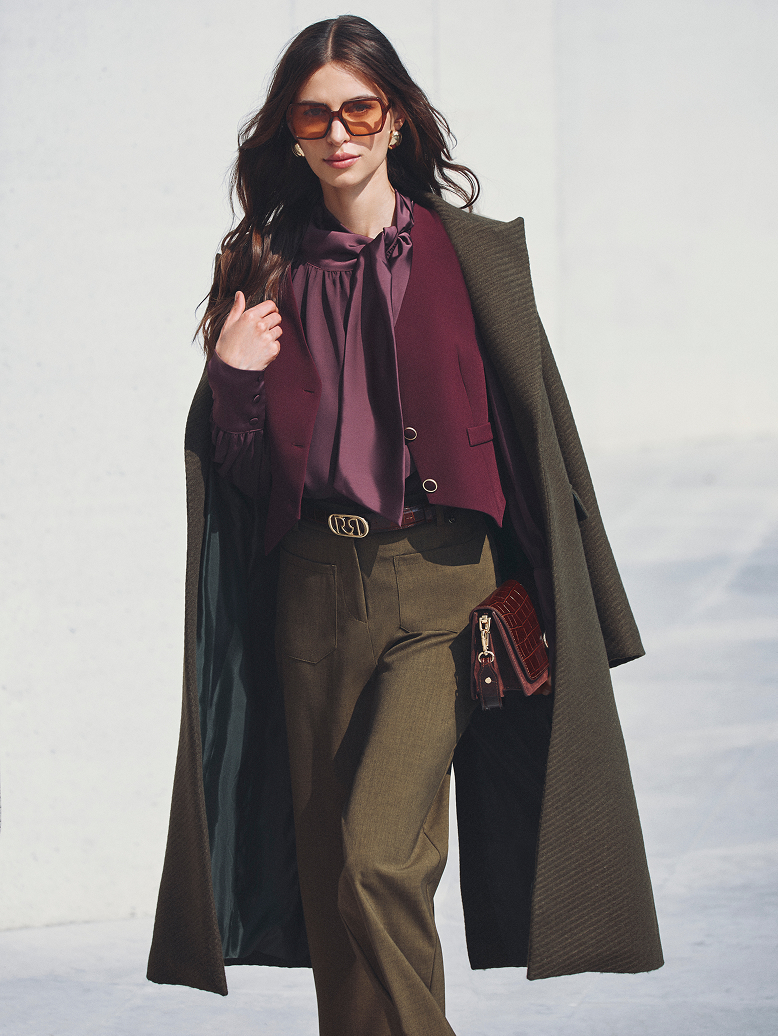 Donna con blusa bordeaux e cappotto verde, in piedi, con uno sfondo naturale